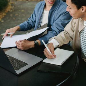 From above of positive male students working on laptop with documents and notebook while doing homework together